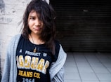 A young person with disheveled hair, wearing a black T-shirt with the text 'A PRODUCT OF JAGUAR JEANS CO. 1964' and a grey open hoodie. The individual is standing against a rough white wall and a dark, tiled floor is visible in the background.