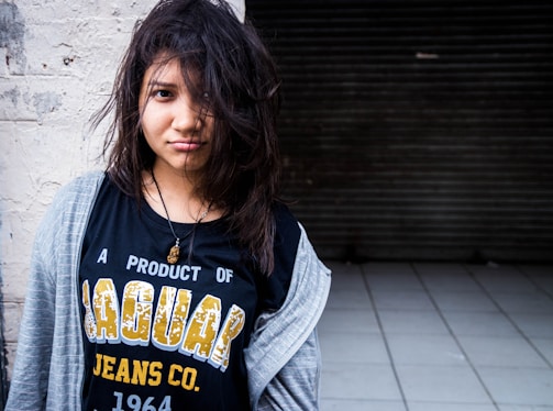 A young person with disheveled hair, wearing a black T-shirt with the text 'A PRODUCT OF JAGUAR JEANS CO. 1964' and a grey open hoodie. The individual is standing against a rough white wall and a dark, tiled floor is visible in the background.