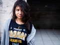 A young person with disheveled hair, wearing a black T-shirt with the text 'A PRODUCT OF JAGUAR JEANS CO. 1964' and a grey open hoodie. The individual is standing against a rough white wall and a dark, tiled floor is visible in the background.