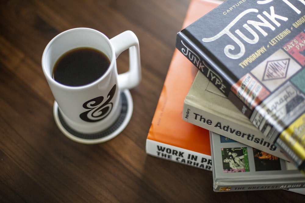 taza de café blanca y negra cerca de una pila de libros sobre una superficie de madera marrón