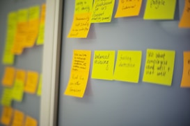 Multiple yellow sticky notes with handwritten text are attached to a glass wall. Some notes contain brainstorming ideas or reminders. The background includes additional sticky notes in various colors, slightly out of focus, suggesting a workspace or collaborative setting.