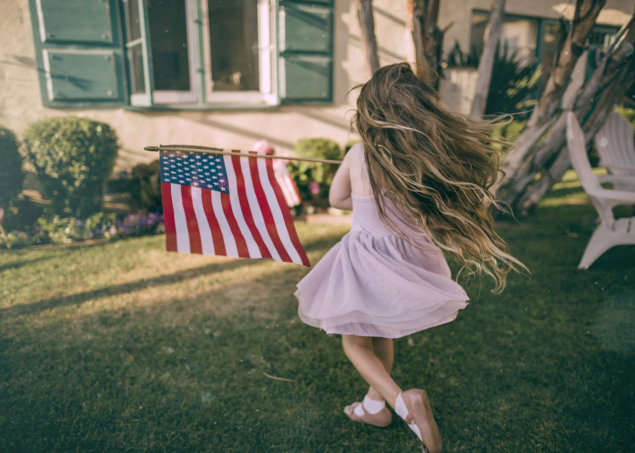A young girl in a pink dress runs across her front lawn carrying a small American flag