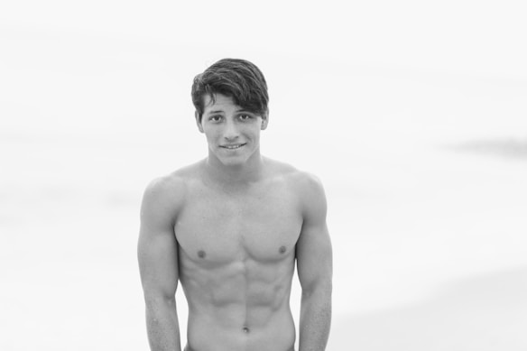 A black and white photograph of a shirtless young man with short, dark hair, standing against a softly blurred background, possibly a beach or open water.