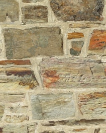 A close-up view of a stone wall with irregularly shaped stones and varying sizes, set in a beige mortar. The stones feature different earthy tones, including browns, grays, and hints of orange and green. The surface has a rough texture with visible layers and grain.