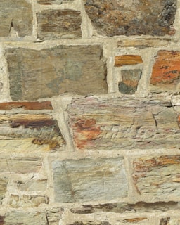 A close-up view of a stone wall with irregularly shaped stones and varying sizes, set in a beige mortar. The stones feature different earthy tones, including browns, grays, and hints of orange and green. The surface has a rough texture with visible layers and grain.