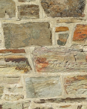 A close-up view of a stone wall with irregularly shaped stones and varying sizes, set in a beige mortar. The stones feature different earthy tones, including browns, grays, and hints of orange and green. The surface has a rough texture with visible layers and grain.