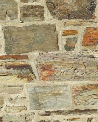 A close-up view of a stone wall with irregularly shaped stones and varying sizes, set in a beige mortar. The stones feature different earthy tones, including browns, grays, and hints of orange and green. The surface has a rough texture with visible layers and grain.
