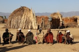 A group of seven people are seated on the ground, each holding a staff, in front of traditional huts made of sticks and straw. The background features a clear sky and distant mountains.