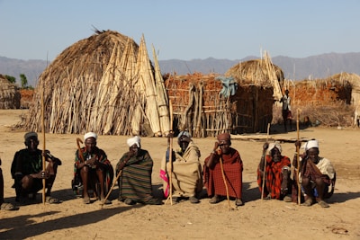 A group of seven people are seated on the ground, each holding a staff, in front of traditional huts made of sticks and straw. The background features a clear sky and distant mountains.