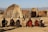 A group of seven people are seated on the ground, each holding a staff, in front of traditional huts made of sticks and straw. The background features a clear sky and distant mountains.
