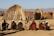 A group of seven people are seated on the ground, each holding a staff, in front of traditional huts made of sticks and straw. The background features a clear sky and distant mountains.