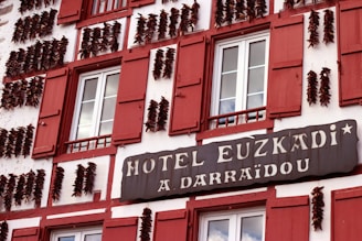 A building facade with red shutters and strings of dried peppers adorning its exterior. The signage reads 'Hotel Euzkadi A. Darraidou'. The windows are symmetrically placed and the overall architecture has a traditional European style.