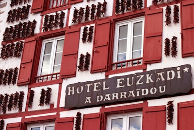 A building facade with red shutters and strings of dried peppers adorning its exterior. The signage reads 'Hotel Euzkadi A. Darraidou'. The windows are symmetrically placed and the overall architecture has a traditional European style.