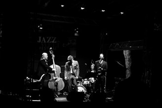Four musicians perform on a dimly lit stage set for a jazz festival, with a double bassist, drummer, saxophonist, and a musician in a hat. The atmosphere is intimate and focused, highlighted by stage lighting that casts shadows around the edges.