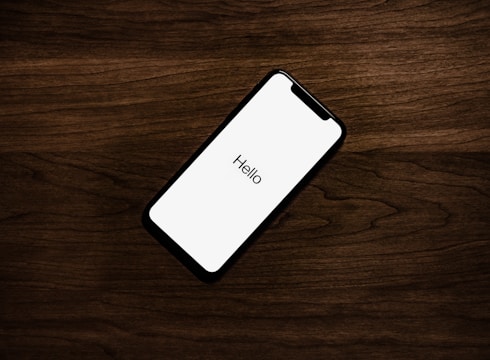 A smartphone with a blank white screen displaying the word 'Hello' is placed on a dark wooden surface. The phone is positioned at an angle, and the lighting emphasizes the texture of the wood.