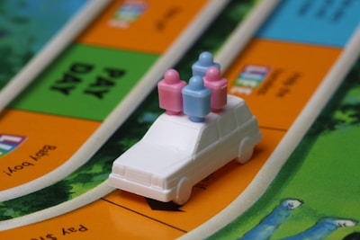 A miniature white car with four small, colorful peg figures on top, situated on a board game with vibrant paths and areas labeled with text. The background includes green, orange, and blue sections representing different parts of the game board.