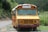A yellow school bus with a weathered exterior is parked on the side of a rural road, surrounded by lush green vegetation and hills in the background. The bus displays some signs of rust and aging, and there are plants growing around the area.