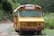 A yellow school bus with a weathered exterior is parked on the side of a rural road, surrounded by lush green vegetation and hills in the background. The bus displays some signs of rust and aging, and there are plants growing around the area.