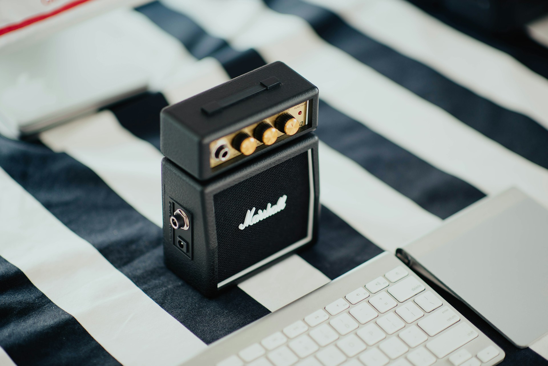 A small, black Marshall amplifier is placed on a striped black and white surface. It is surrounded by a white keyboard and a closed laptop, suggesting a blend of musical and digital elements.