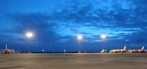 The image depicts an airport tarmac during dusk with several airplanes parked and illuminated by bright floodlights. The sky is a mix of deep blue and hints of cloud cover.