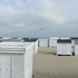 A series of white beach cabins are lined up on sandy terrain under a cloudy sky. A red and white checkered flag is visible, indicating the wind direction. The setting appears to be a calm coastal area with footprints leading between the cabins.