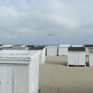 A series of white beach cabins are lined up on sandy terrain under a cloudy sky. A red and white checkered flag is visible, indicating the wind direction. The setting appears to be a calm coastal area with footprints leading between the cabins.