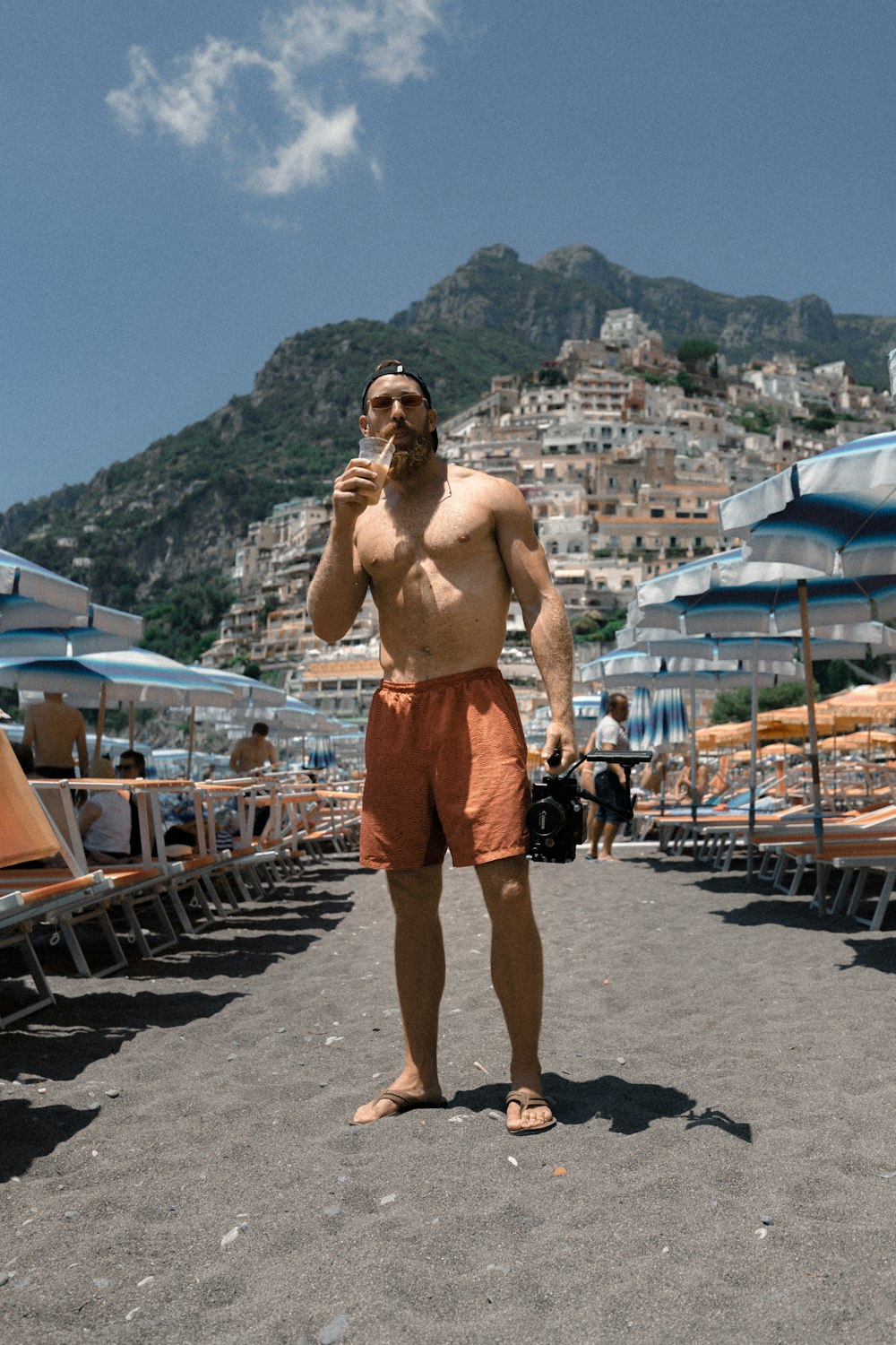 homem vestindo shorts laranja em pé na areia da praia enquanto bebe no copo