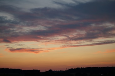 A sunset scene with large, sweeping clouds painted in shades of pink and purple, set against an orange and yellow horizon. The silhouette of a distant landscape, including trees and hills, is visible at the bottom.