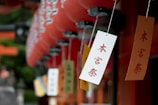 Several colorful paper tags with Japanese characters hang from strings, accompanied by rows of red paper lanterns in the background, creating a vibrant and cultural setting.