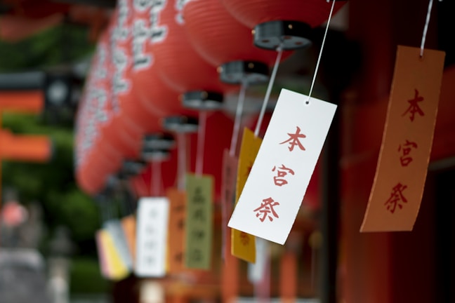 Several colorful paper tags with Japanese characters hang from strings, accompanied by rows of red paper lanterns in the background, creating a vibrant and cultural setting.