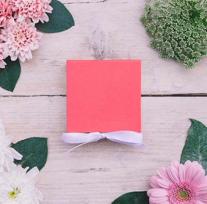 A flat-lay arrangement featuring a pinkish-red square box tied with a white ribbon, surrounded by an assortment of flowers and green leaves on a light wooden background.
