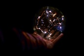 A hand gently cradles a transparent sphere filled with small, glowing lights against a dark background. The lights form delicate patterns resembling a constellation or network inside the orb, casting a warm glow.