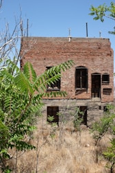 A two-story abandoned brick building with boarded-up windows surrounded by overgrown vegetation. The structure appears weathered and neglected, with dry grass covering the foreground.