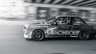 A high-speed car, adorned with racing decals, speeds along a road with a dynamic motion blur effect, emphasizing its velocity. The driver, wearing a helmet, is focused and in full racing gear. The background features an urban setting under a bridge, adding to the sense of motion.