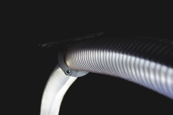 A coiled metal hose with a shiny, reflective surface extends from a dark background. The focus is on the curved section of the hose, revealing its detailed ribbed texture and metallic material.