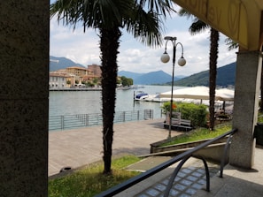 A serene lakeside scene with moored boats and a view of distant mountains under a partly cloudy sky. Palm trees line the walkway leading to the water, and there are outdoor seating areas with large canopies. The area has a peaceful and inviting ambiance.