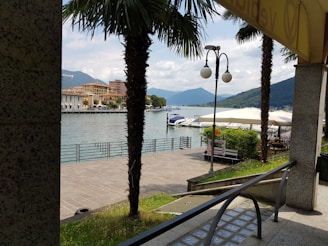 A serene lakeside scene with moored boats and a view of distant mountains under a partly cloudy sky. Palm trees line the walkway leading to the water, and there are outdoor seating areas with large canopies. The area has a peaceful and inviting ambiance.