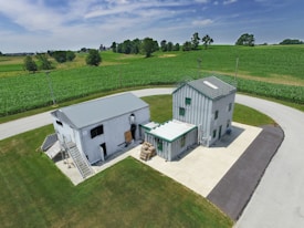 Two small industrial-style buildings with metal roofs are situated on a concrete lot surrounded by lush green fields. A country road loops around the lot. A few trees are scattered in the background under a partly cloudy sky.