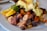 A plate of food featuring colorful roasted potatoes, slices of oranges, cherry tomatoes, and a dish topped with creamy sauce and greens. The potatoes exhibit a variety of colors including purple, pink, and orange. The meal is garnished with fresh green herbs.
