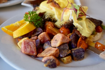 A plate of food featuring colorful roasted potatoes, slices of oranges, cherry tomatoes, and a dish topped with creamy sauce and greens. The potatoes exhibit a variety of colors including purple, pink, and orange. The meal is garnished with fresh green herbs.