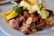 A plate of food featuring colorful roasted potatoes, slices of oranges, cherry tomatoes, and a dish topped with creamy sauce and greens. The potatoes exhibit a variety of colors including purple, pink, and orange. The meal is garnished with fresh green herbs.