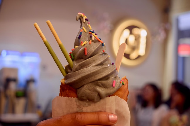 A hand holds a dessert featuring soft-serve ice cream with a grayish hue, topped with multicolored sprinkles. The ice cream is nestled in a pastry cone and paired with a few Pocky sticks. The background is slightly blurred with warm lighting, hinting at an indoor setting, possibly a cafe or dessert shop.