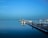 A tranquil marina scene with several sailboats and small boats anchored at docks. The water is calm, reflecting the vessels and the clear blue sky. Seagulls can be seen flying subtly in the distance.