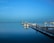 A tranquil marina scene with several sailboats and small boats anchored at docks. The water is calm, reflecting the vessels and the clear blue sky. Seagulls can be seen flying subtly in the distance.
