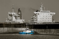 A large cargo ship with a prominent superstructure is docked in still waters, with a small blue tugboat maneuvering alongside it. The vessel's hull is dark, while the upper structures are white, and a plume of smoke rises from one of the smokestacks.