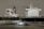 A large cargo ship with a prominent superstructure is docked in still waters, with a small blue tugboat maneuvering alongside it. The vessel's hull is dark, while the upper structures are white, and a plume of smoke rises from one of the smokestacks.