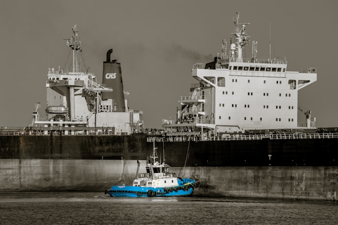 A large cargo ship with a prominent superstructure is docked in still waters, with a small blue tugboat maneuvering alongside it. The vessel's hull is dark, while the upper structures are white, and a plume of smoke rises from one of the smokestacks.