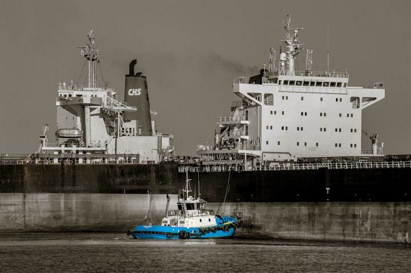 A large cargo ship with a prominent superstructure is docked in still waters, with a small blue tugboat maneuvering alongside it. The vessel's hull is dark, while the upper structures are white, and a plume of smoke rises from one of the smokestacks.