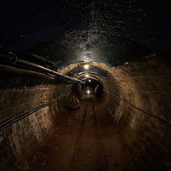 A dimly lit underground tunnel with brick walls and a rough, dirt-covered floor. Overhead lights create a series of illuminated spots along the curved ceiling. Pipes and cables run along the walls, contributing to the industrial atmosphere.