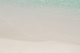 Gentle waves of clear turquoise water wash onto a smooth, white sandy beach. The sand has a soft, fine texture, appearing almost untouched.